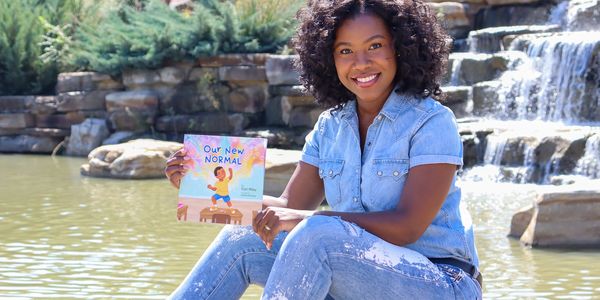 Cori Riley, author of "Our New Normal", holding a copy of the book, sitting in front of a waterfall