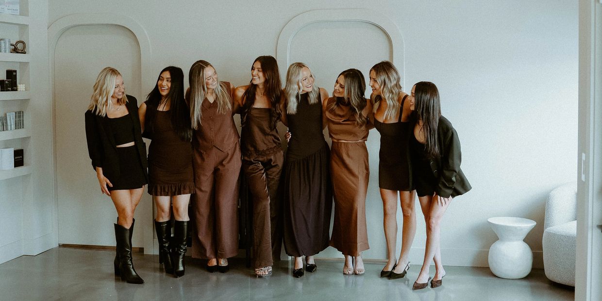 Eight women in brown and black outfits smiling and posing indoors.