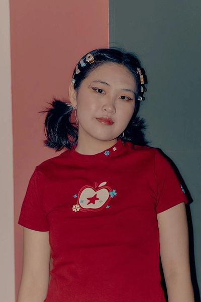 Performer ENHUI stands against a pink and grey background, wearing a red t-shirt and playful haircli