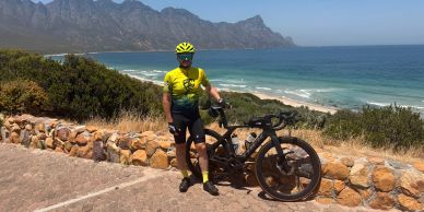 Cyclist posing with bike near a coastal road with mountains and ocean.