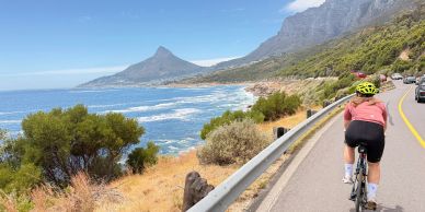 Cyclist rides along a coastal road with mountains and ocean views on a sunny day.