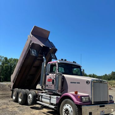 A purple and white dump truck unloading gravel under a clear blue sky.