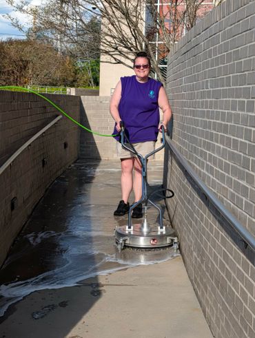 Woman pressure washing a concrete walkway near a brick wall.