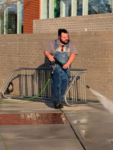 Man power washing a sidewalk near a brick building.