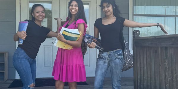 Three young women smiling and holding notebooks outside a house.
