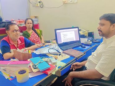 Three people engaged in a discussion at a cluttered office desk with a computer screen on.
