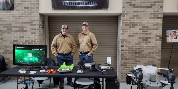 Two men in matching shirts stand behind a tech display table with drones.