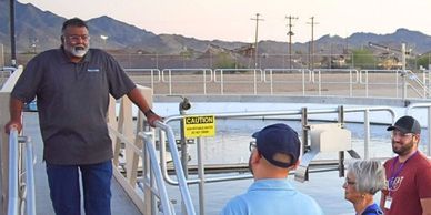 A man speaks to a small group near a waterfront dock with mountains in the background.