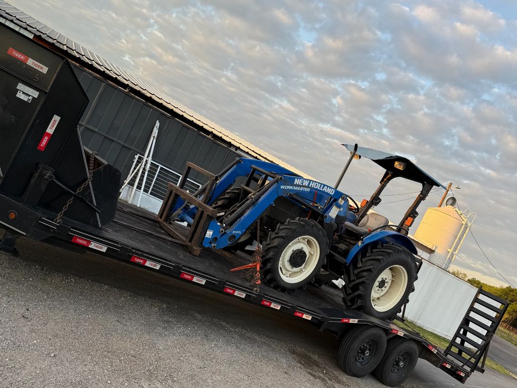 Blue New Holland Workmaster tractor on a flatbed trailer outdoors.