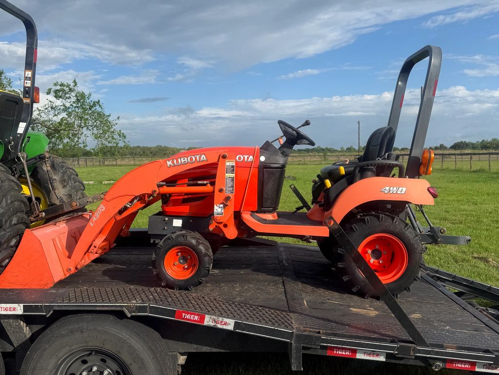 Orange Kubota tractor with front loader on a trailer under a partly cloudy sky.