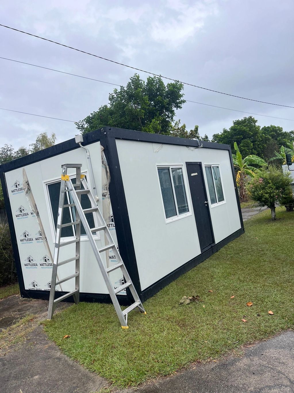 A tilted modular building on grass with a ladder leaning against it.
