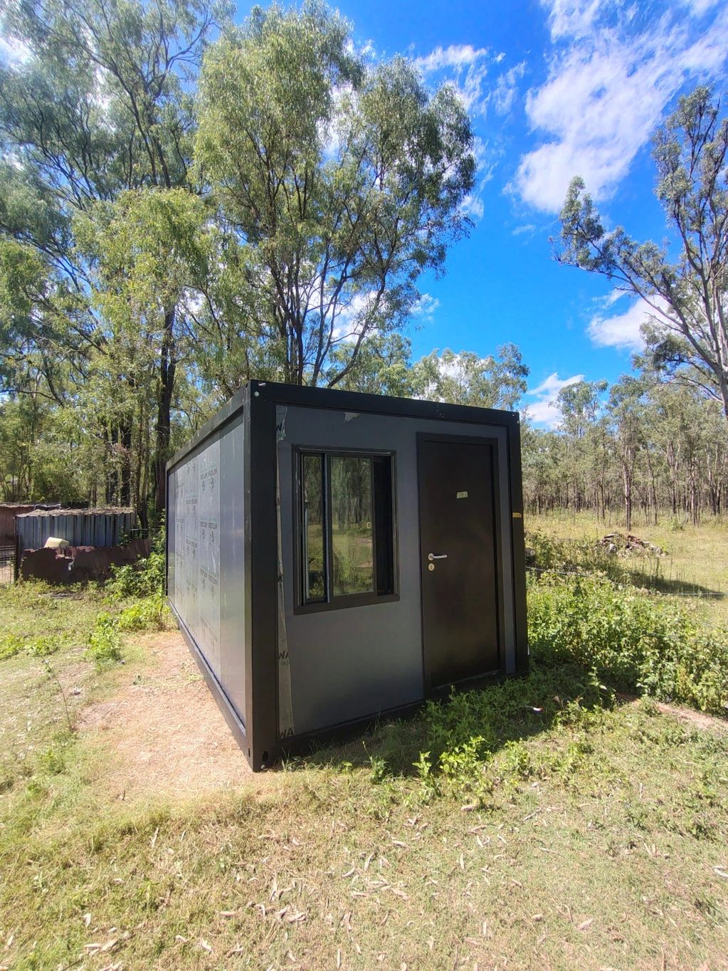 A small black container cabin in a grassy outdoor area with trees.