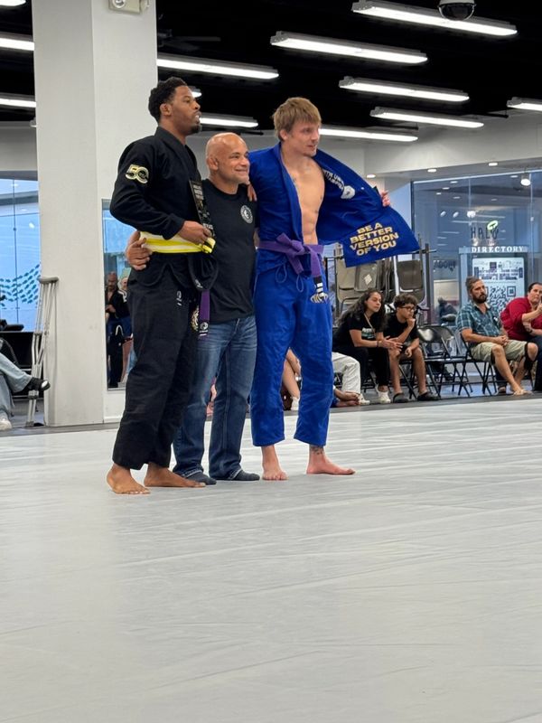Three men posing on a martial arts mat, two in gis and one in casual clothes.