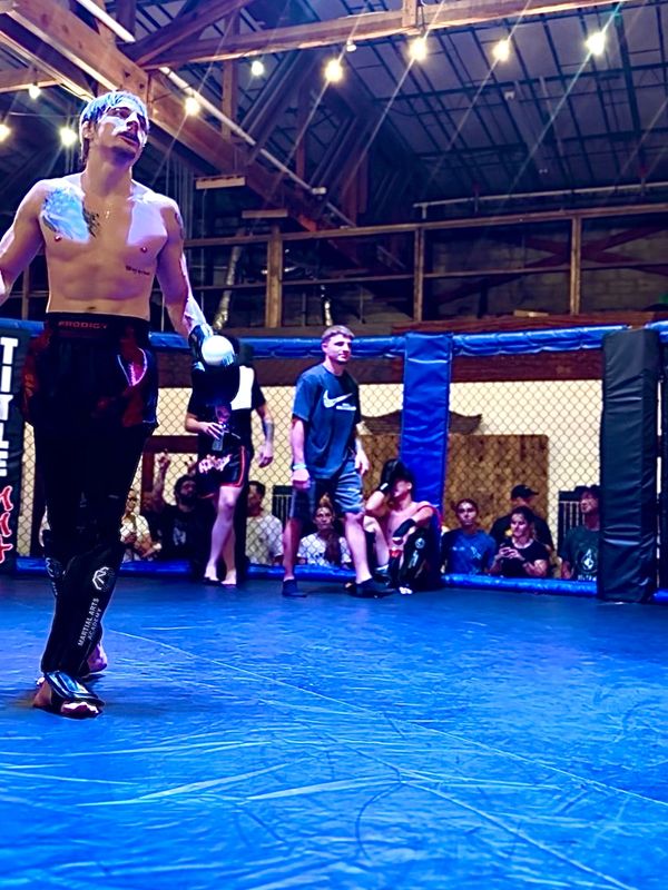 A shirtless fighter walks confidently in a cage ring with an audience watching.