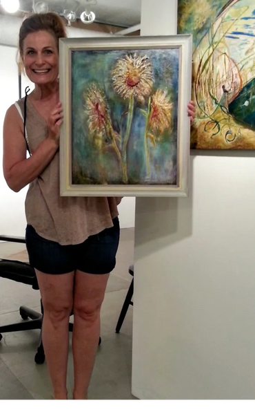 a smiling woman holding up a painting of three while dandelions