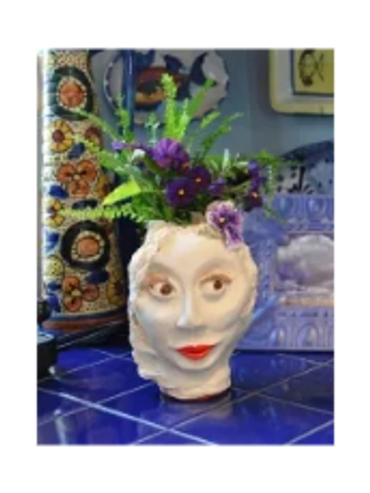 A hand-sculpted clay vase of a woman's face, containing purple and green floral arrangement.