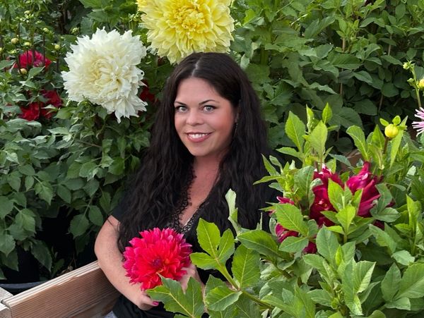 A woman with dark hair smiles holding a vibrant pink flower among large yellow and white blooms.