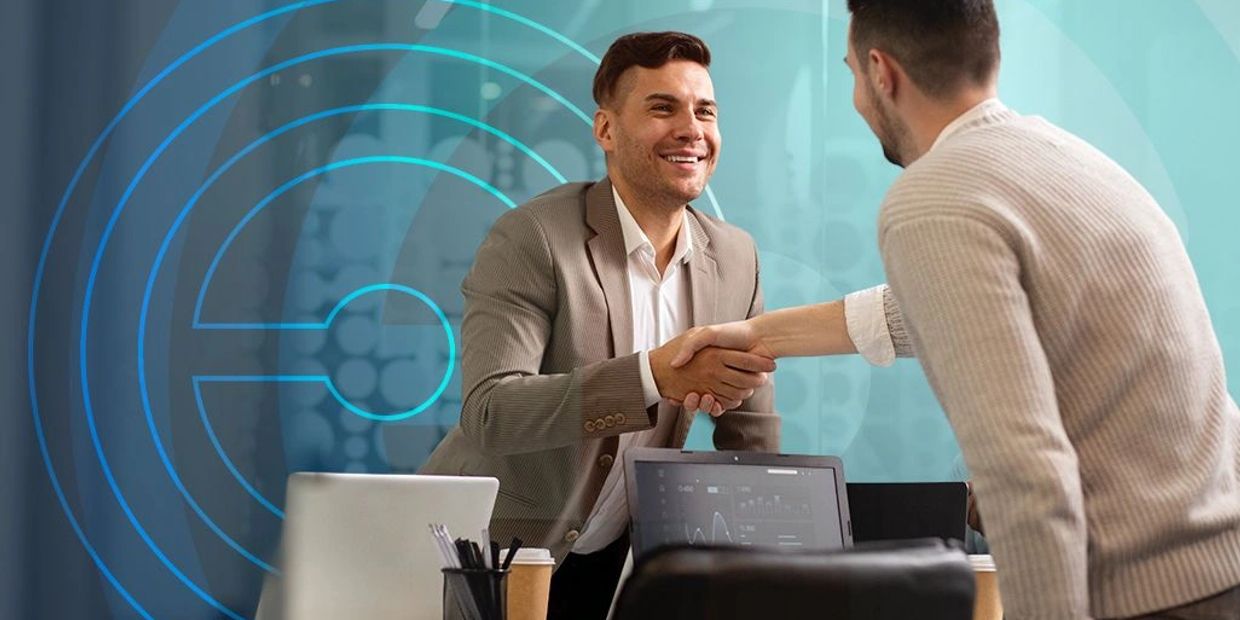 Two men shaking hands in a business setting with laptops on the table.