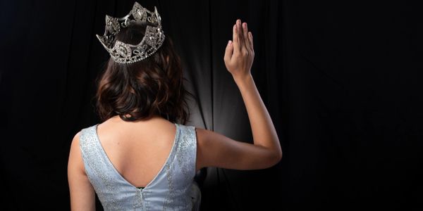 Woman in a crown and dress raising her hand against a black backdrop.