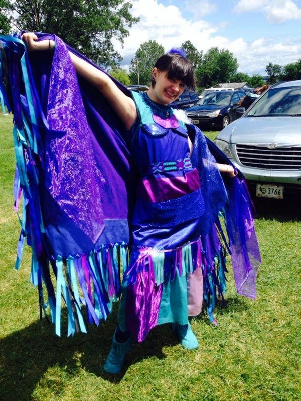 A native woman fancy shawl dancer that is wearing bright blue. There are cars in the background.