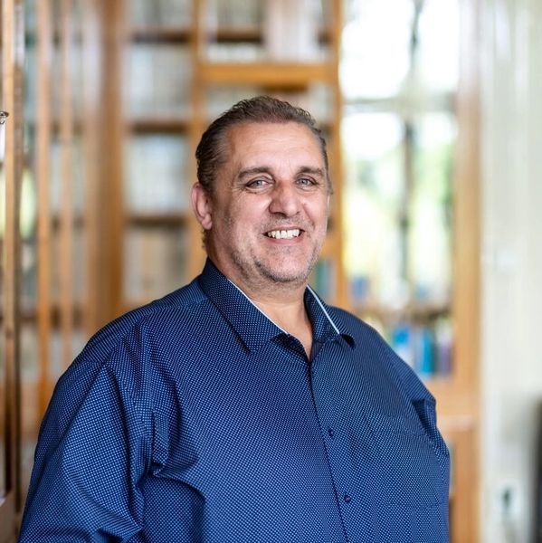 Smiling man in a blue shirt standing indoors with blurred background.
