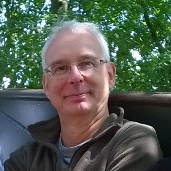 Smiling elderly man wearing glasses outdoors with green trees in the background.