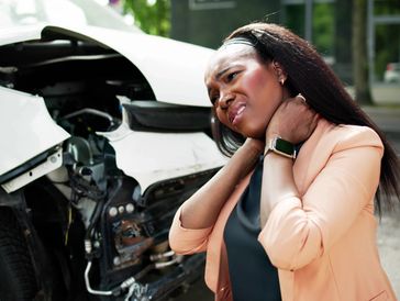 A woman in a peach blazer stands near a damaged white car, visibly distressed