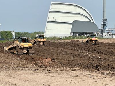 Three bulldozers working on soil near a large industrial building.