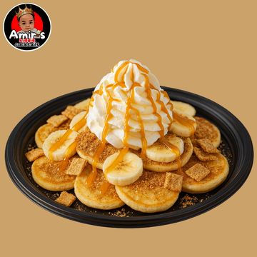 Mini pancakes topped with whipped cream, banana slices, caramel drizzle, and crunchy cereal on a black plate.