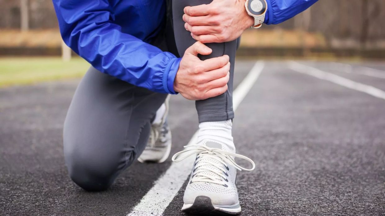 Runner holding injured leg on track, wearing blue jacket and gray pants.