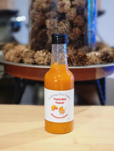 A bright orange bottle of Ladybug Hot Sauce on a wooden surface with pinecones in the background.