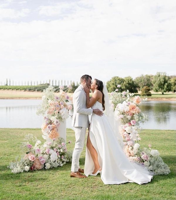 Bride and groom share a kiss at an outdoor lakeside wedding ceremony adorned with pastel flowers.