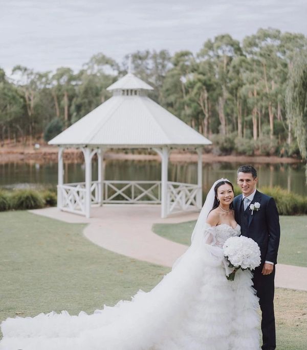 Happy bride and groom posing outdoors in front of a white gazebo by the water.
