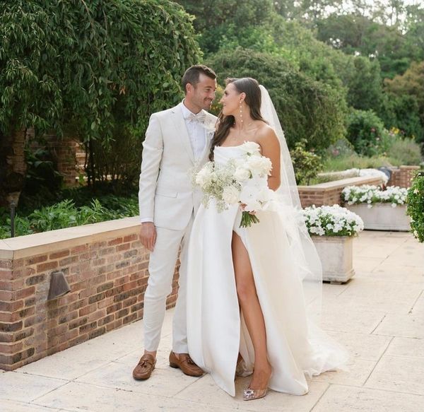 A bride and groom in elegant white attire share a loving gaze outdoors.