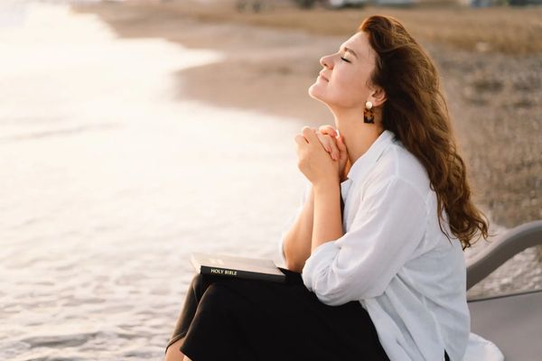 A young woman holding a Bible to her chest and praying outdoors by a river at sunset.