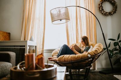 A woman reading a book in a cozy round chair by a window with warm lighting.