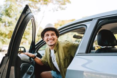 A young man smiling while getting out of a car on a sunny day.