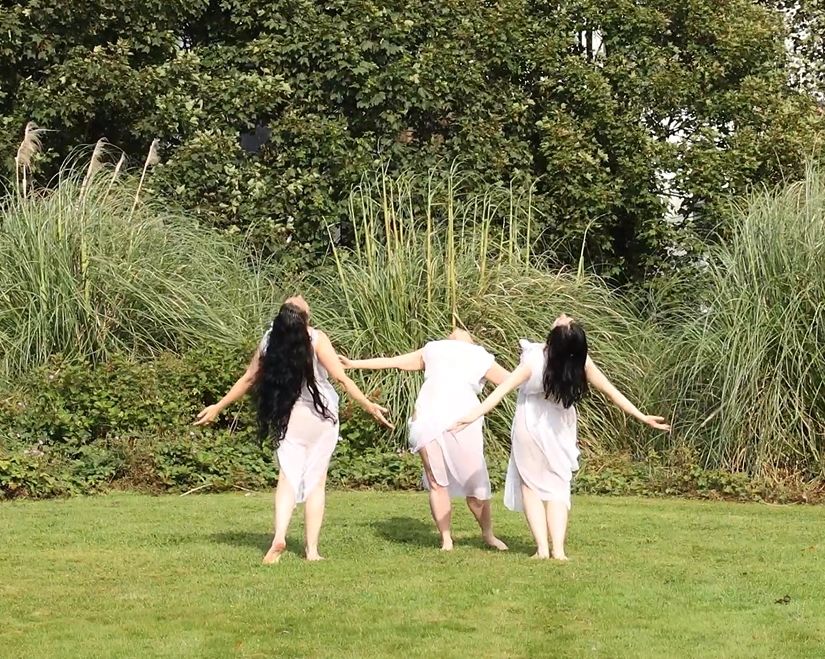 Three women in white dresses dancing outdoors with arms outstretched.