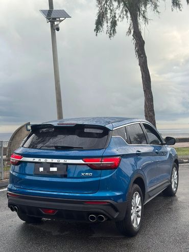 Blue Proton X50 SUV parked on a wet road under cloudy skies.