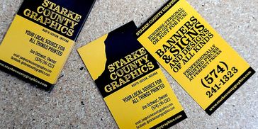 Three business cards scattered on a desk that promote Starke County Graphics. Black and yellow.