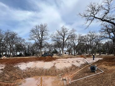 Construction site with muddy ground, scattered pipes, and leafless trees under a cloudy sky.