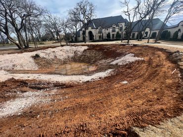 Excavation site for a pond near a large house with bare trees.