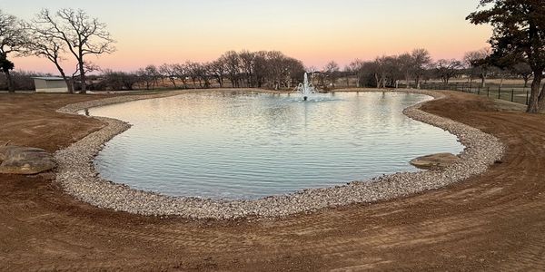 A serene pond with a central fountain at sunset, surrounded by bare trees and a gravel border.