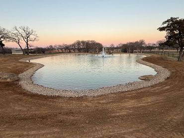 A serene pond with a fountain surrounded by bare trees at sunset.