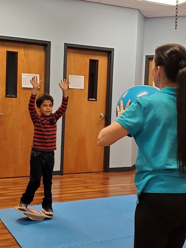 Therapist throwing ball at child who is balancing on a balance board. Occupational Therapy