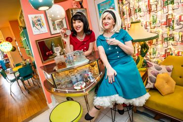 Two women in retro dresses enjoying drinks in a colorful vintage living room.