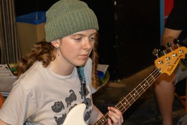 Young woman playing a white Fender bass guitar, wearing a green knit hat.