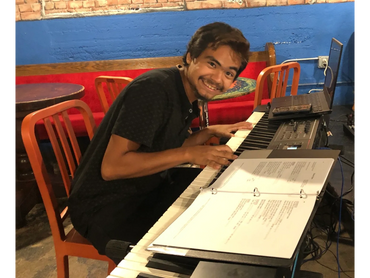 Young man happily playing keyboard with sheet music and laptop nearby.