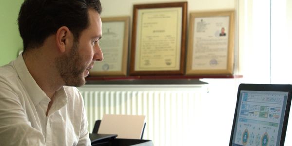 Man in white shirt working on a laptop in an office with framed certificates.