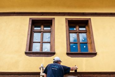 Man on a ladder applying wood stain to the window trim.